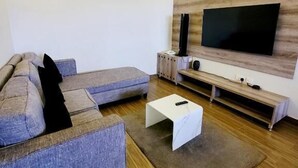 A modern living room with a sectional sofa, a flat-screen TV mounted on a wooden wall, a white coffee table, and a wooden floor.