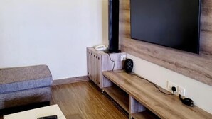 A modern living room with a flat-screen TV mounted on a wooden wall, a grey sofa, a white ottoman, and a wooden bench.