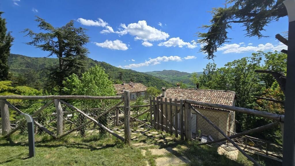 A rustic wooden fence leading to a building with a tiled roof, surrounded by lush greenery and hills in the background.