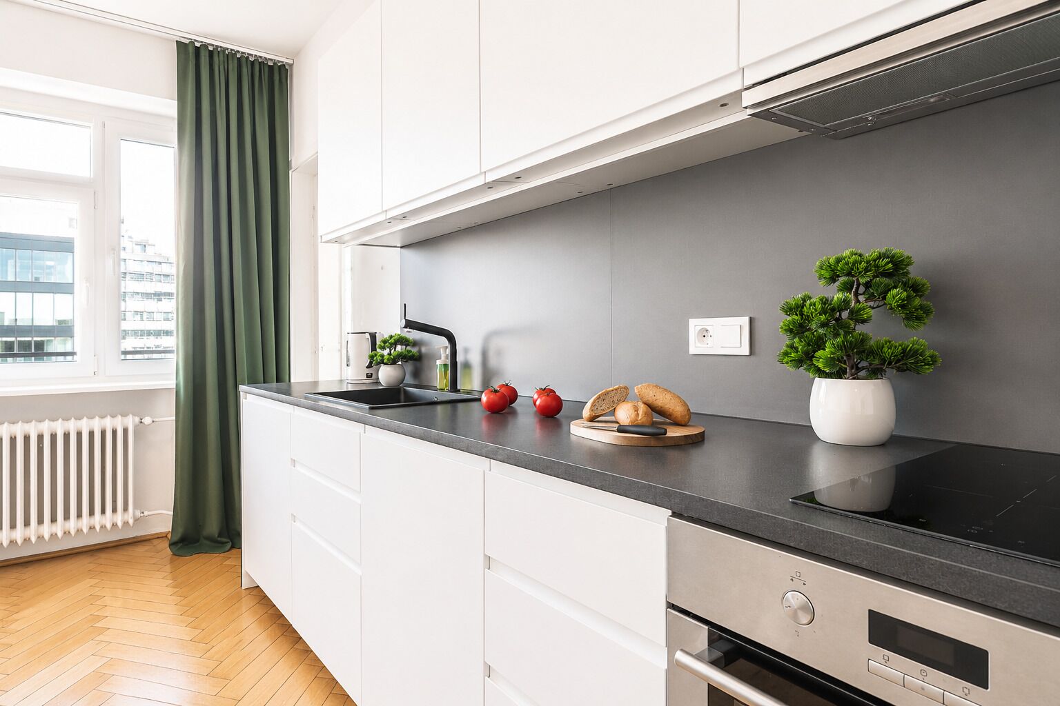 Una cocina moderna con encimera negra, armarios blancos, horno integrado y ventana con vistas a edificios.