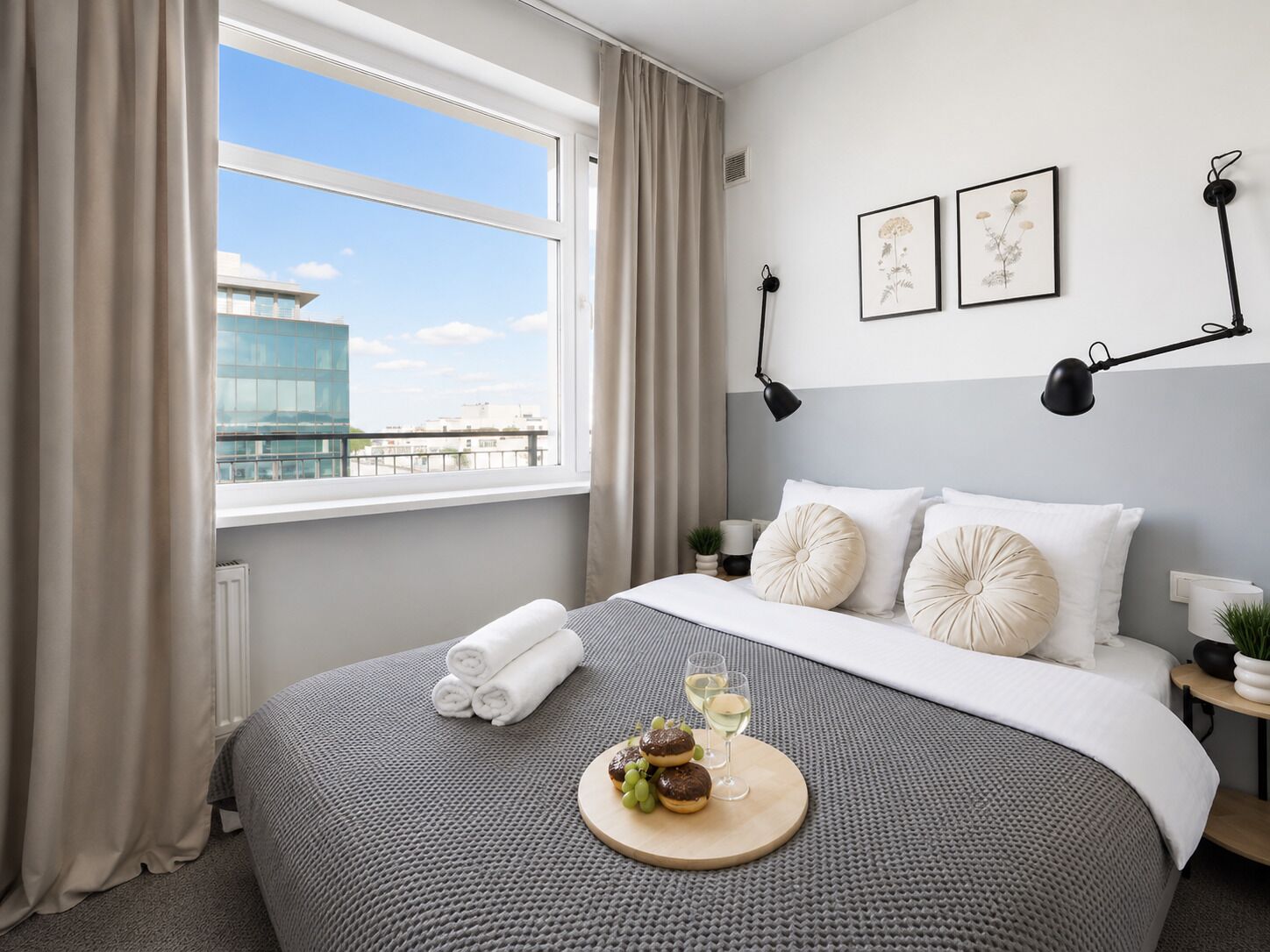 A modern hotel room with a large bed, a view of a building through the window, and two wall-mounted lamps.