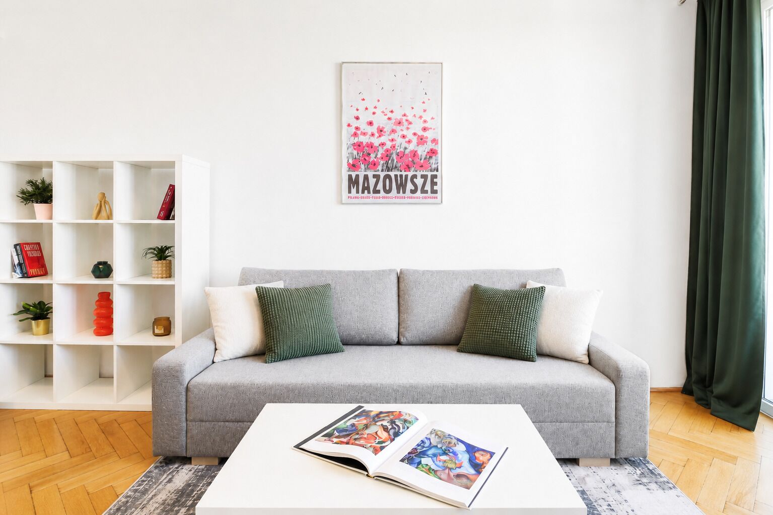 Ein Wohnzimmer mit einem grauen Sofa, einem weißen Couchtisch mit einer Zeitschrift, einer weißen Regaleinheit mit Dekoration und einer Wand mit einem gerahmten Bild.
