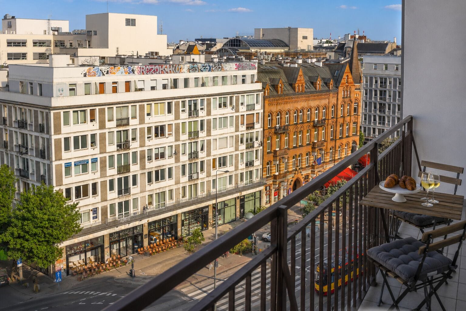 Eine Stadtansicht mit einer Mischung aus modernen und historischen Gebäuden, einem Balkon mit Tisch und Stuhl sowie einer Straßenbahn.