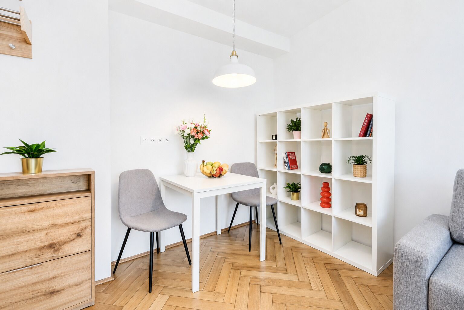 A modern living room with a white dining table, two grey chairs, a white bookshelf, and a herringbone-patterned wooden floor.