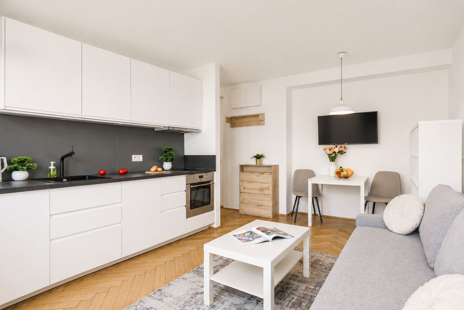 Una cocina moderna con armarios blancos, una encimera negra y un horno. Hay un comedor con una mesa y sillas blancas, y un sofá gris con un cojín blanco.