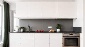 A modern kitchen with white cabinets, a black countertop, a built-in oven, and a window with curtains. - Lumina 2-room Apart with Balcony Center (Warszawa)