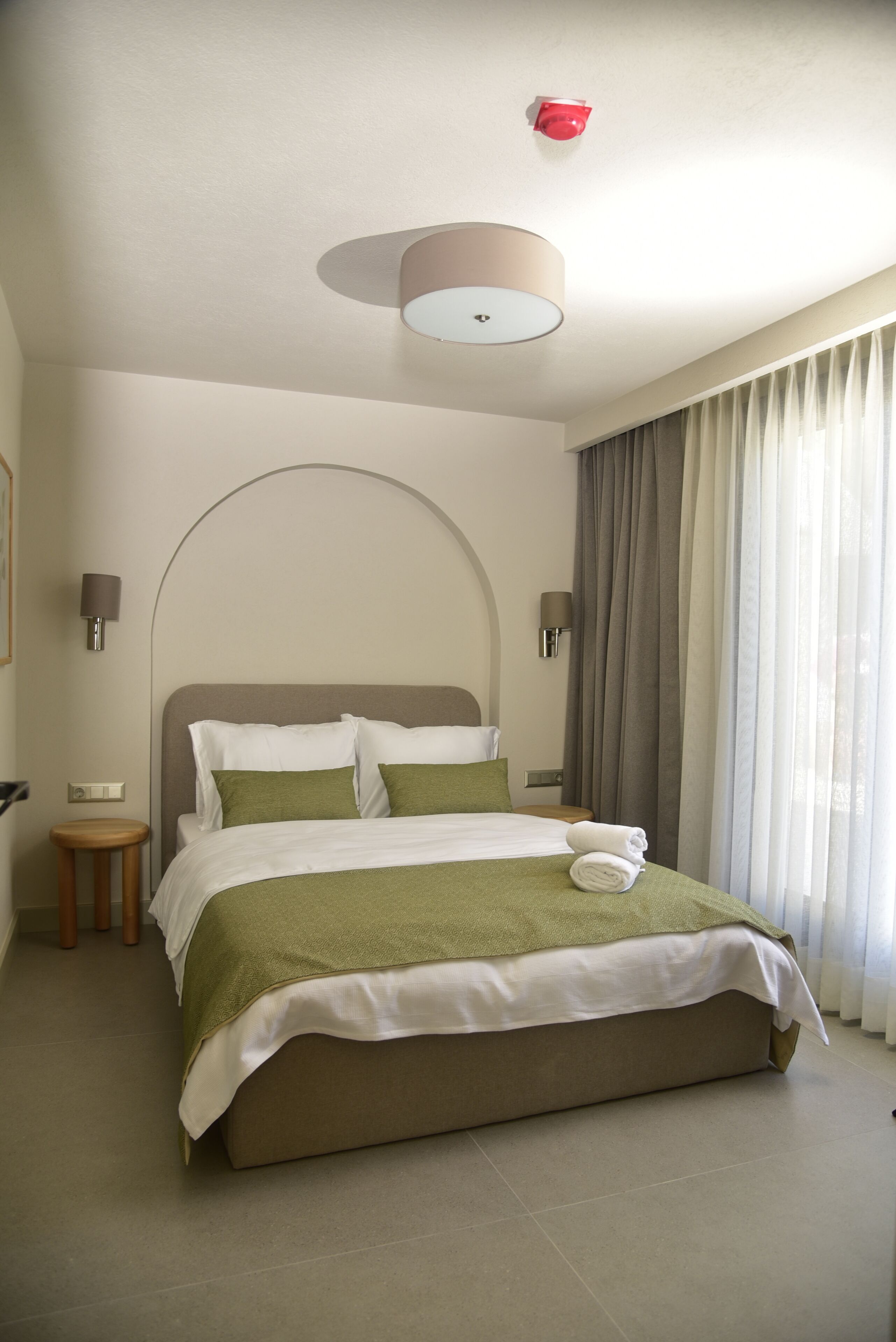 Uma cama bem arrumada com colcha verde e branca, uma luminária redonda no teto e uma luz de emergência vermelha.