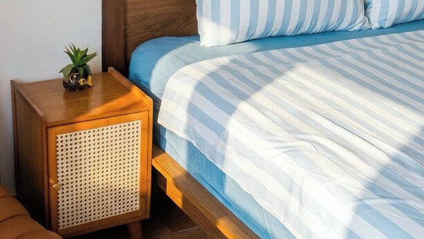 A bed with striped bedding, a wooden headboard, a bedside table with a rattan speaker, and a brown sofa.