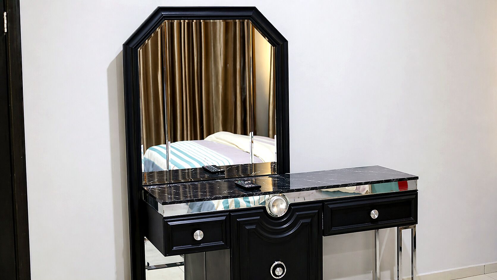 A black vanity with a mirror, a bed with striped bedding, and a red object on the bed.