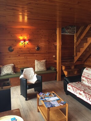 A cozy cabin interior with a wooden wall, a staircase, a sofa, a chair, and a coffee table with magazines. - 100 M² DUPLEX "SPECIAL OFFER" IN THE HEART OF LES CARROZ—Sleeps 9, 3 bedrooms, 2 bathrooms (Arâche -La-Frasse)