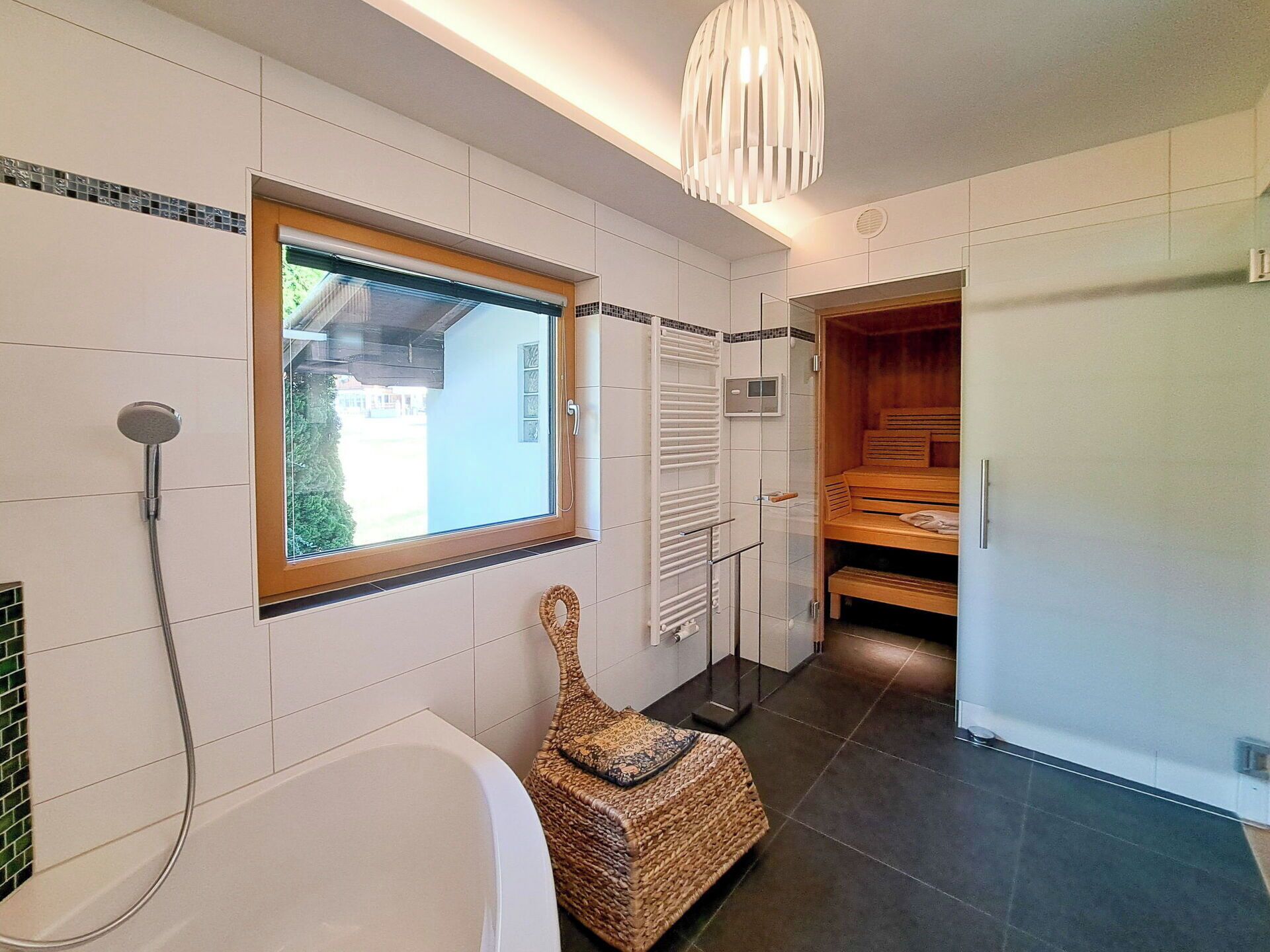 A modern bathroom with a bathtub, a shower, a wooden bench, a woven stool, and a window with a view.