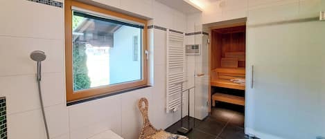 A modern bathroom with a bathtub, a shower, a wooden bench, a woven stool, and a window with a view.