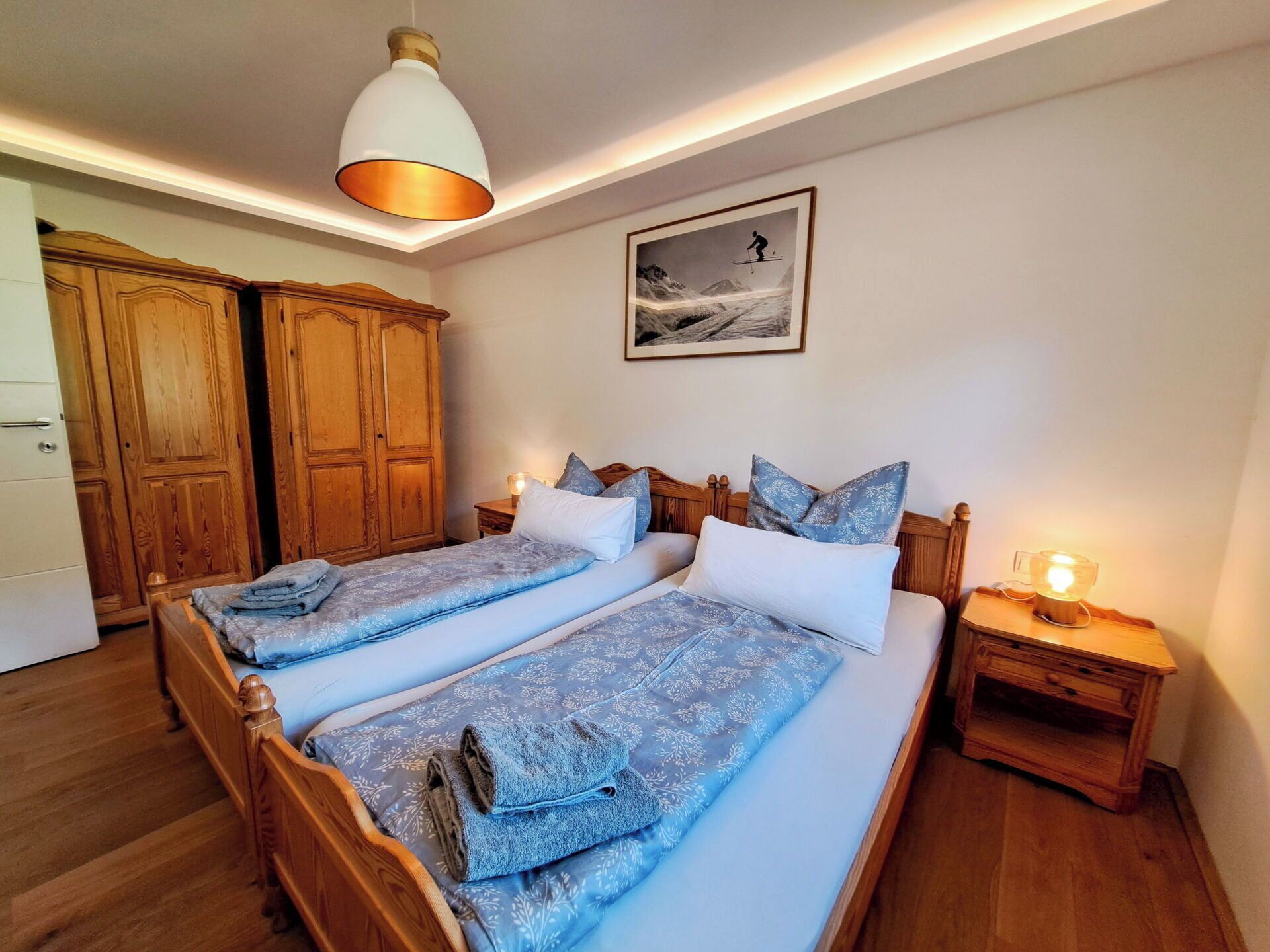 A double bed with patterned bedding, a wooden wardrobe, a framed picture on the wall, and a bedside table with a lamp.