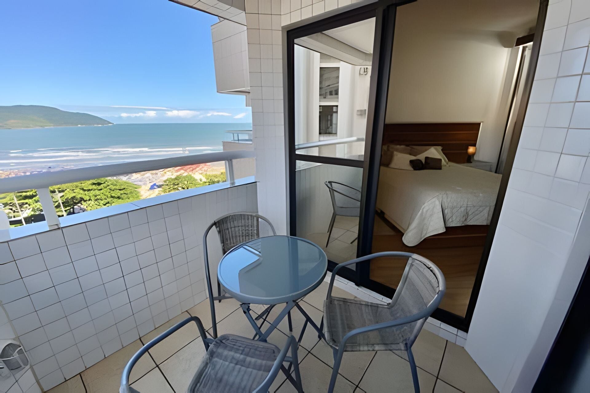 A balcony with a glass table and chairs, a bed with a wooden headboard, and a view of the ocean.