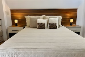 Una cama bien hecha con edredón blanco, dos mesitas de noche con lámparas y un cabecero de madera.