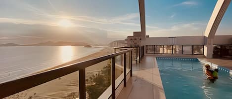 A modern hotel with a pool, a balcony with a view of the beach, and a clear sky.