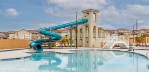 Ein Schwimmbecken mit einer großen blauen Rutsche, einer weißen Treppe und einem beige gefärbten Gebäude mit Rundbogenfenstern.