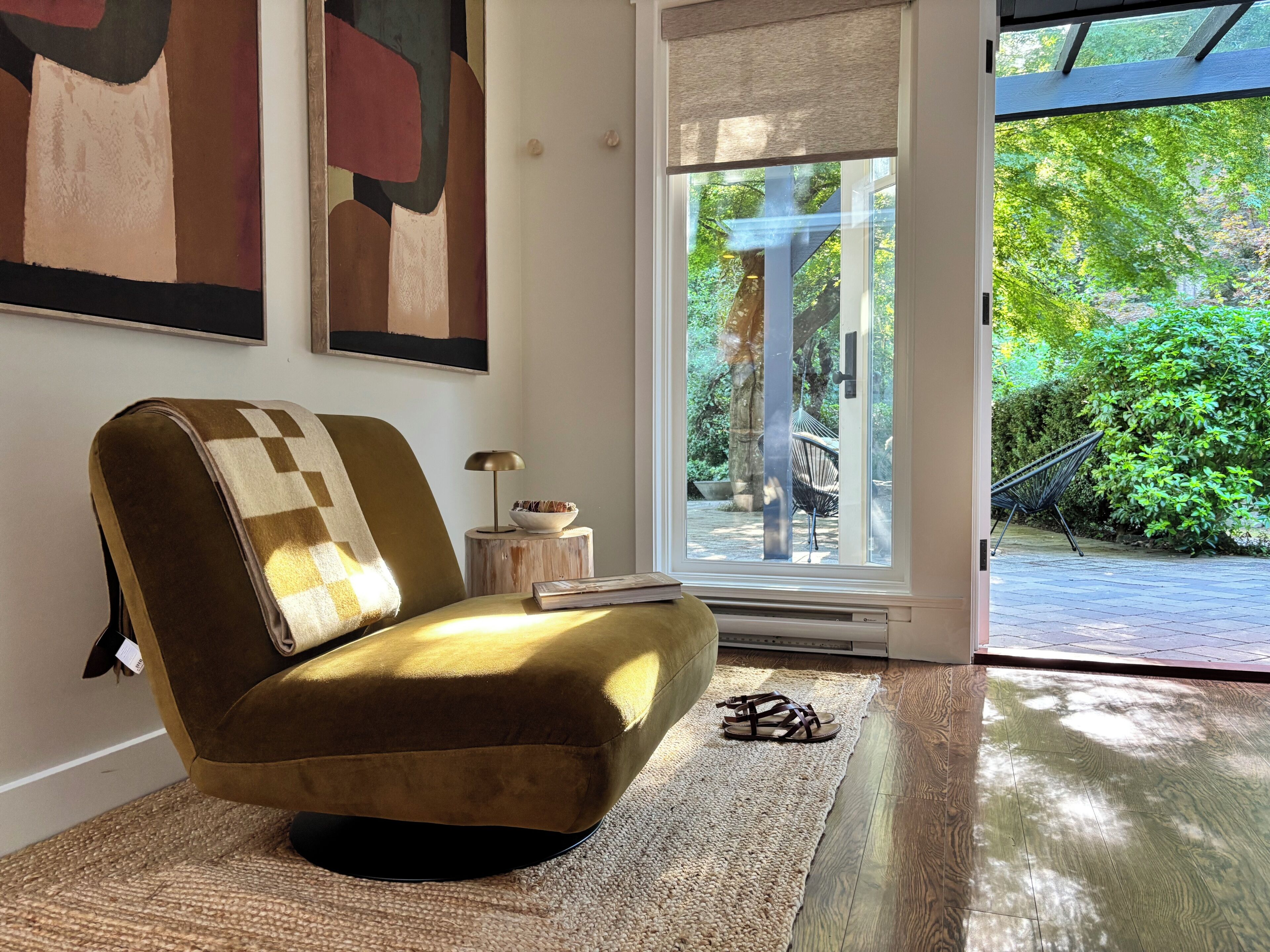 Ein Zimmer mit bequemem Ohrensessel, einem kleinen Tisch mit Lampe und Blick auf einen Garten durch eine Glastür.