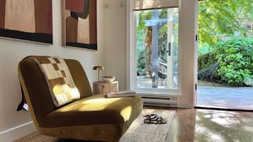 Ein Zimmer mit bequemem Ohrensessel, einem kleinen Tisch mit Lampe und Blick auf einen Garten durch eine Glastür.