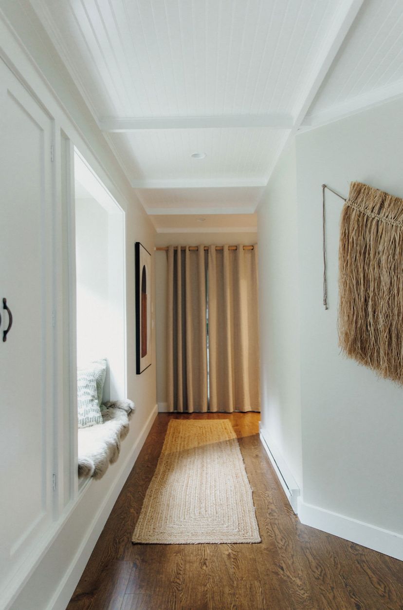 A hallway with wooden flooring, a beige rug, a framed picture on the wall, and a curtain at the end.