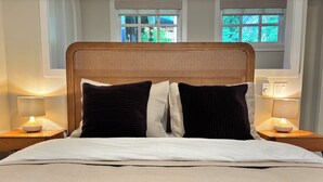 A bed with a woven headboard, two bedside lamps, and a view of greenery through windows.