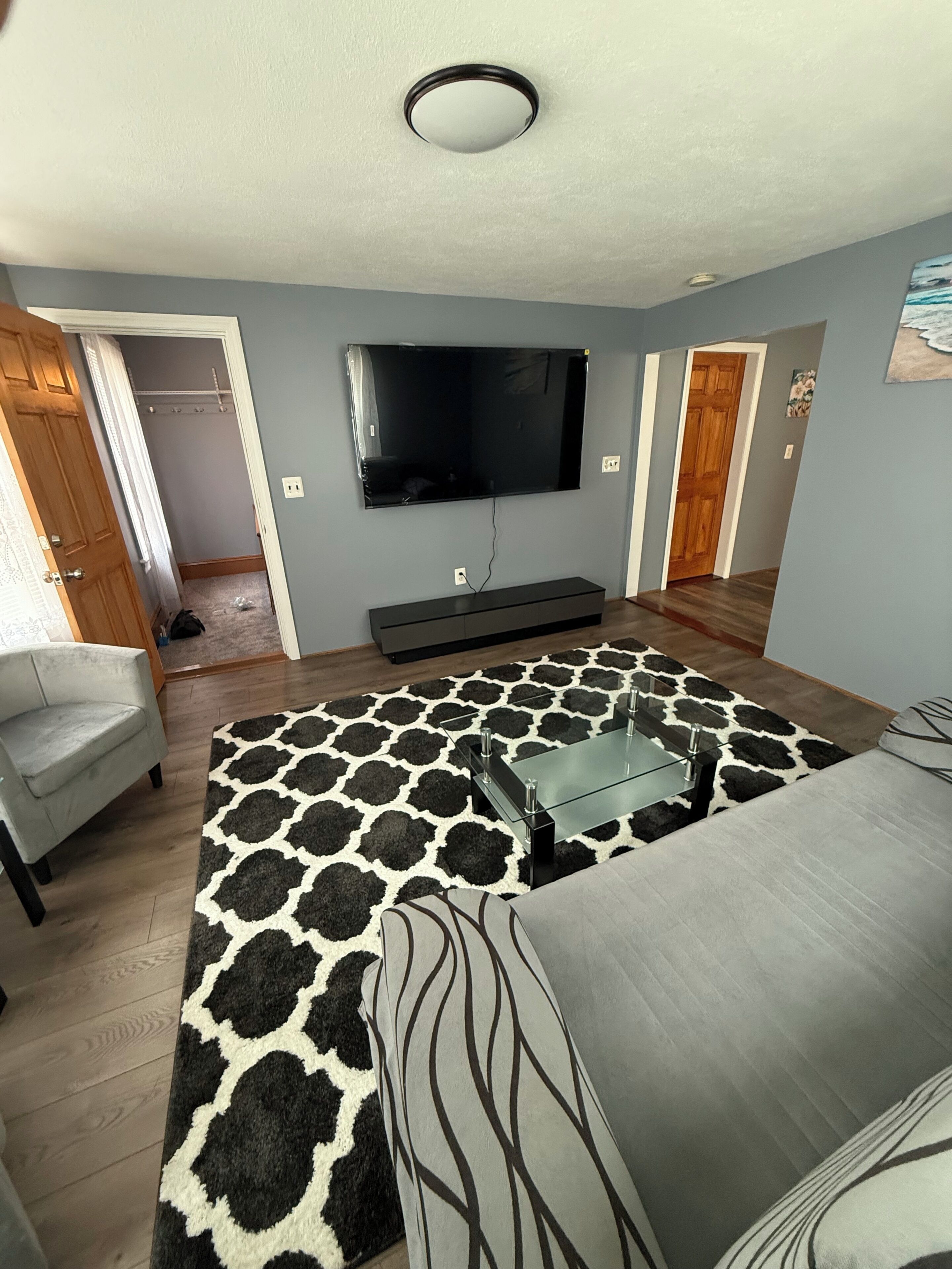A living room with a flat-screen TV mounted on the wall, a glass coffee table, a patterned rug, a grey sofa, and a wooden floor.
