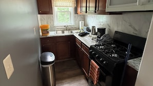 A compact kitchen with wooden cabinets, a stainless steel sink, a microwave, a stove, and a trash bin.