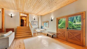 Un intérieur spacieux avec un escalier en bois, un coin salon, un bureau et une grande porte en bois donnant sur un paysage verdoyant.