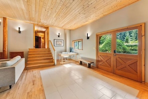 A spacious interior with a wooden staircase, a seating area, a desk, and a large wooden door with a view of greenery.