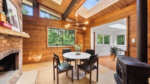 Une pièce chaleureuse avec une cheminée en briques, des poutres apparentes en bois au plafond, une table ronde entourée de chaises et de grandes fenêtres donnant sur l’extérieur.