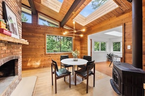 A cozy room with a brick fireplace, wooden ceiling beams, a round table with chairs, and large windows offering a view of the outdoors.