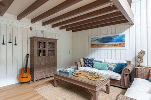 A room with a wooden ceiling, a white wall with a painting, a sofa with patterned cushions, a coffee table with books and baskets, and a guitar against the wall. - Villa chunga piscine chauffee jacuzzi et clim (Lège-Cap-Ferret)