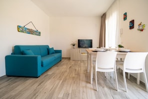 A modern living room with a teal sofa, a dining table with white chairs, a TV, and a wooden floor. - GuestHost - Borgo 84 Marzamemi - Happy Family (Pachino)