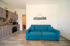 A modern kitchen with wooden cabinets, a teal sofa, and a wall-mounted artwork. - GuestHost - Borgo 84 Marzamemi - Happy Family (Pachino)