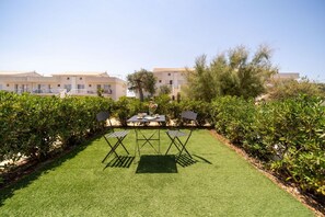 A well-maintained lawn with a table and chairs, surrounded by dense shrubbery and a building in the background. - GuestHost - Borgo 84 Marzamemi - Happy Family (Pachino)