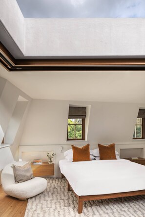 Une chambre à coucher avec un plafond en pente, un lit, une chaise, une table de chevet et deux fenêtres avec des rideaux.