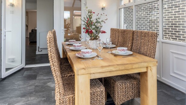 A dining area with a wooden table and woven chairs, a glass enclosure, and a brick wall. - 4-Bedroom Home | Sleeps 10 | Parking | Garden - Pass the Keys (Basildon)