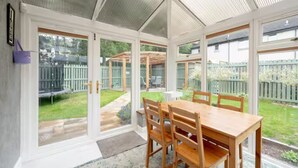 A conservatory with a wooden dining table and chairs, large glass doors leading to a garden. - Modern Family House | Sleeps 5 | Free Parking - Pass the Keys (Edinburgh)