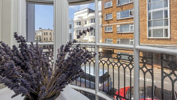 A view from a window with a balcony, overlooking a street with parked cars and buildings. - Charming Georgian Retreat | Walk to Beach & City - Pass the Keys (Brighton)