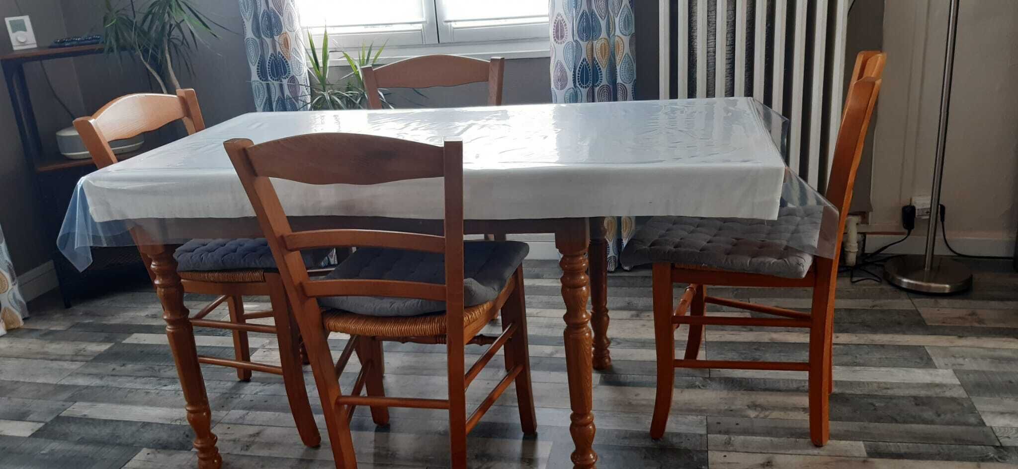 Una mesa de comedor con cuatro sillas de madera, un mantel blanco y una cortina estampada al fondo.