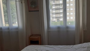 Um quarto com uma cama, um banquinho de madeira e uma janela com vista para um edifício.