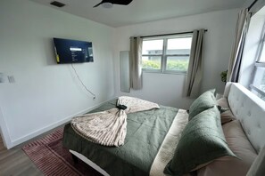 Ein Schlafzimmer mit einem Bett, einem an der Wand montierten Fernseher, einem Fenster mit Vorhängen und Blick ins Grüne.