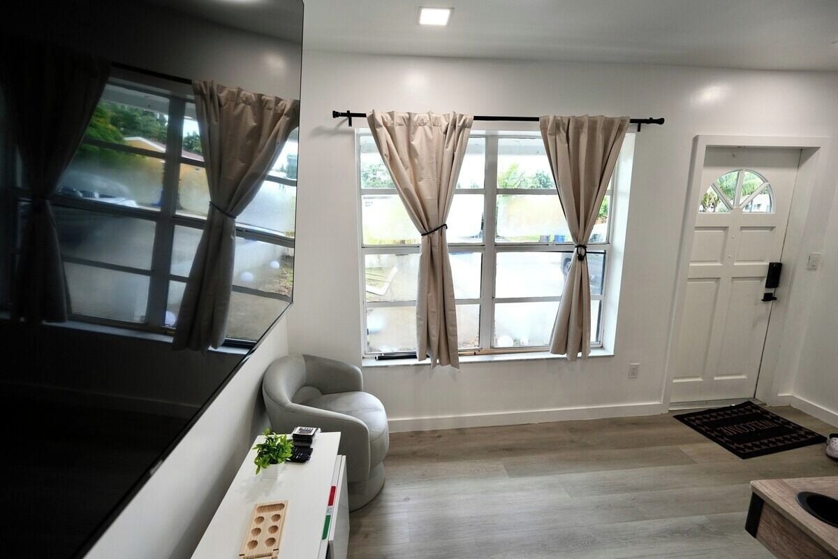 A modern living room with a sofa, a coffee table, and a TV. There are two windows with curtains, a door, and a small rug.
