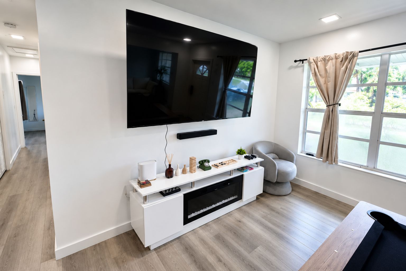 Een moderne woonkamer met een flatscreen televisie aan de muur, een witte tv-meubel met diverse spullen, een grijze fauteuil en een raam met gordijnen.