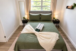 Un lit bien fait, recouvert d’une couverture verte, avec deux oreillers et un plaid blanc. Deux tables de chevet, ornées de lampes et de petites plantes, se trouvent de part et d’autre du lit.