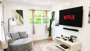 A living room with a round grey sofa, a flat-screen TV mounted on the wall, a potted cactus, and a bookshelf with books and decorative items. - Near Hard Rock . Game Room + 15 Min to Beach (Hollywood)