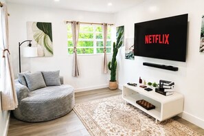 A living room with a round grey sofa, a flat-screen TV mounted on the wall, a potted cactus, and a bookshelf with books and decorative items. - Near Hard Rock . Game Room + 15 Min to Beach (Hollywood)