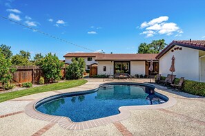A residential backyard with a swimming pool, a patio with outdoor furniture, and a house with a tiled roof.