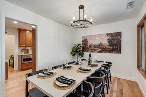 A dining area with a long table set for six, a chandelier, a painting on the wall, and a kitchen in the background.