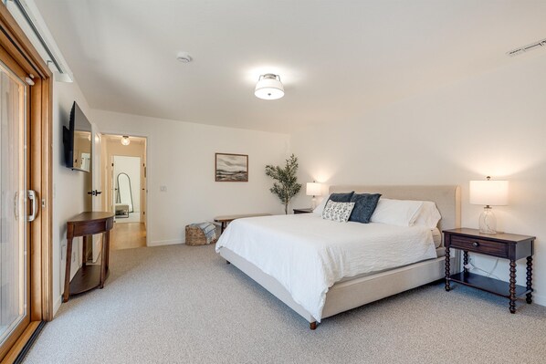 A bedroom with a large bed, bedside tables, a mirror, a wall-mounted TV, and a door leading to another room.